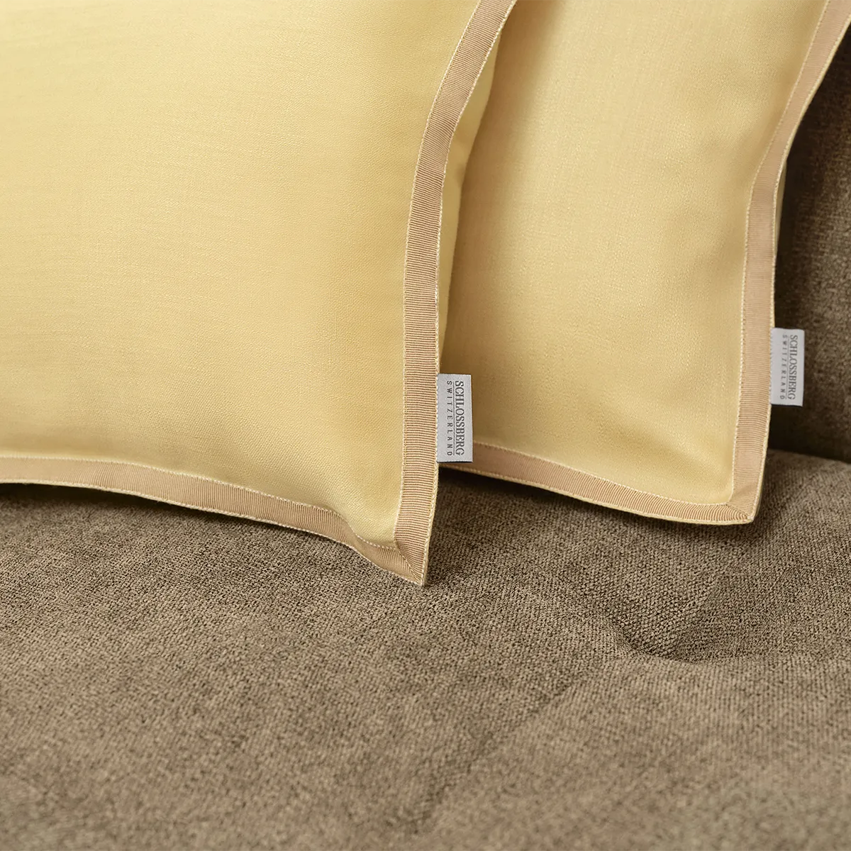 Schlossberg Zierkissen Solene Sun – Edle Verarbeitung im Detail Nahaufnahme von zwei Schlossberg Zierkissen Solene Sun auf einem beigen Sofa. Die feine Paspelierung und der hochwertige Stoff in warmem Gelb unterstreichen die luxuriöse Verarbeitung und sorgen für eine stilvolle Wohnatmosphäre.