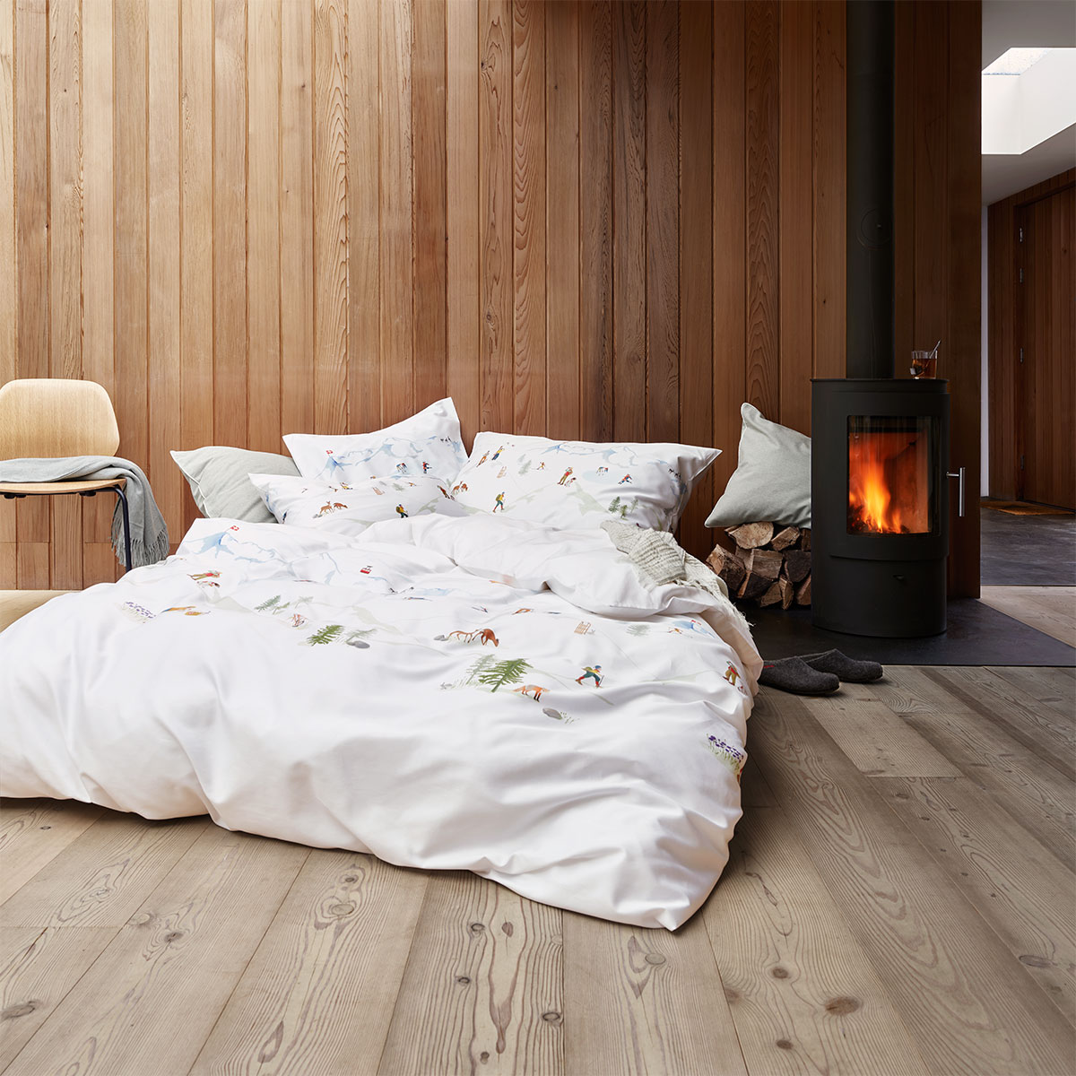Ein gemütlicher Raum mit einem Bett, das mit weisser Bettwäsche und alpinem Motiv bezogen ist. Neben dem Bett steht ein Kaminofen.
