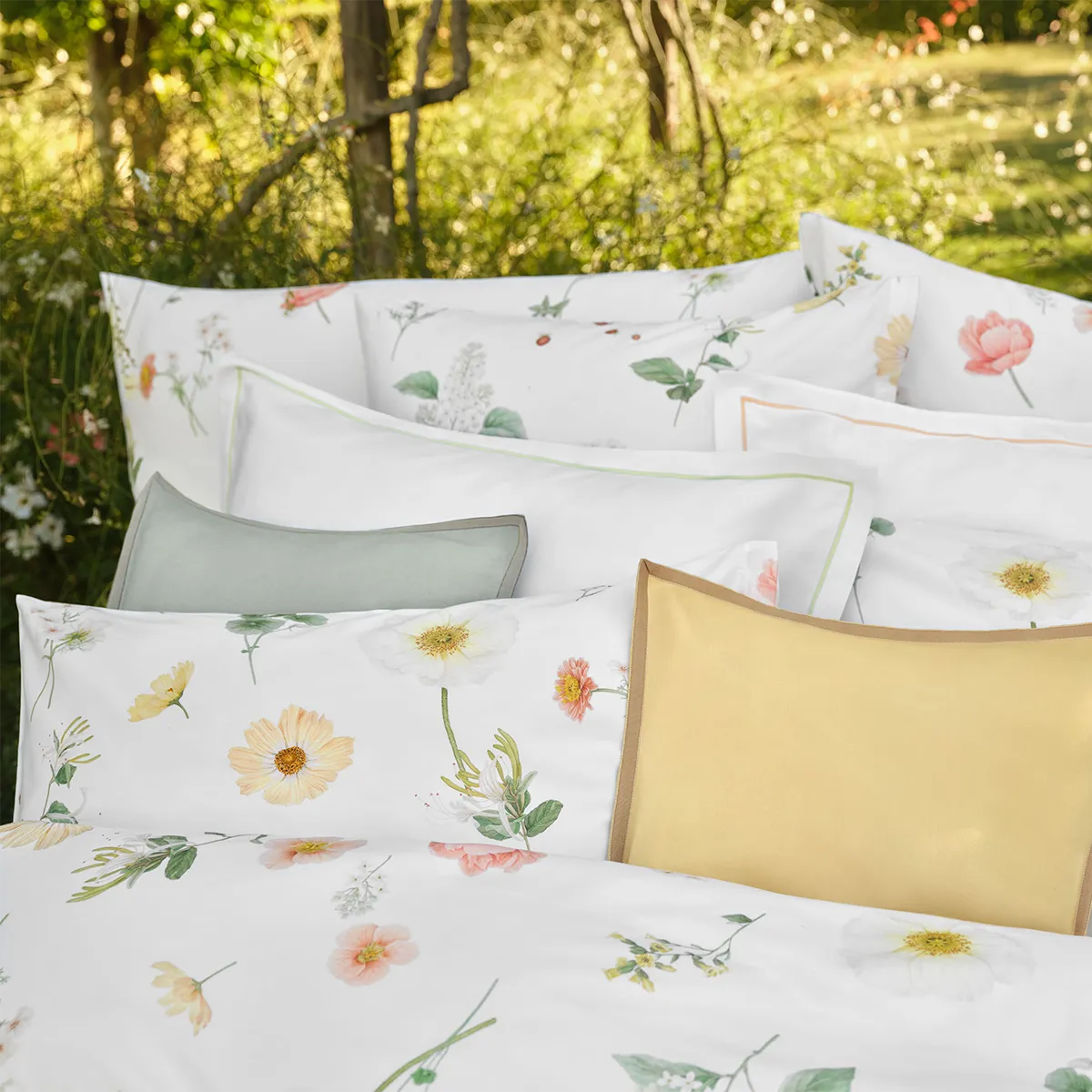 Schlossberg Bettwäsche Zinnia – Solene Kissen im Detail Detailaufnahme von verschiedenen Kissen der Schlossberg Bettwäsche Zinnia, darunter das Solene Kissen in gelb mit braunem Rand, umgeben von weiteren Kissen mit floralen Mustern auf weißem Hintergrund.