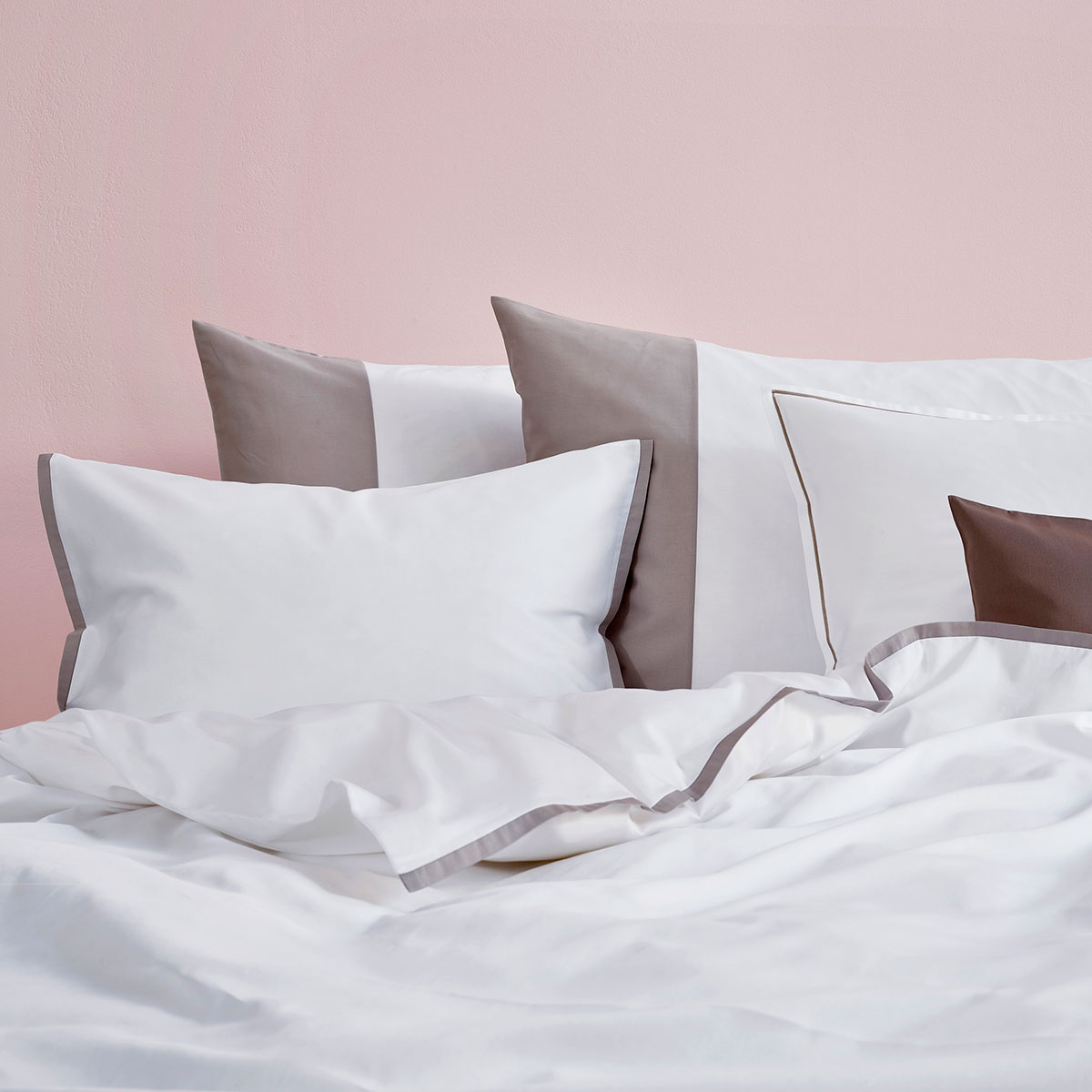 Ein Bett mit weisser und grauer Bettwäsche vor einer rosa Wand. Die Kissen sind in verschiedenen Grössen und Farben, hauptsächlich weiss und grau, dekorativ angeordnet.