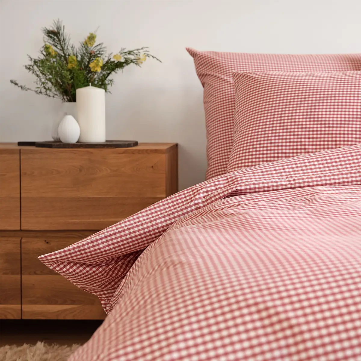 Eine Nahaufnahme der Schlossberg Bettwäsche Andrin Rouge im Schlafzimmer, das mit einem warmen Holzsideboard und einer Vase mit frischen Blumen dekoriert ist. Das rote Karomuster der Bettwäsche verleiht dem Raum eine gemütliche und einladende Atmosphäre