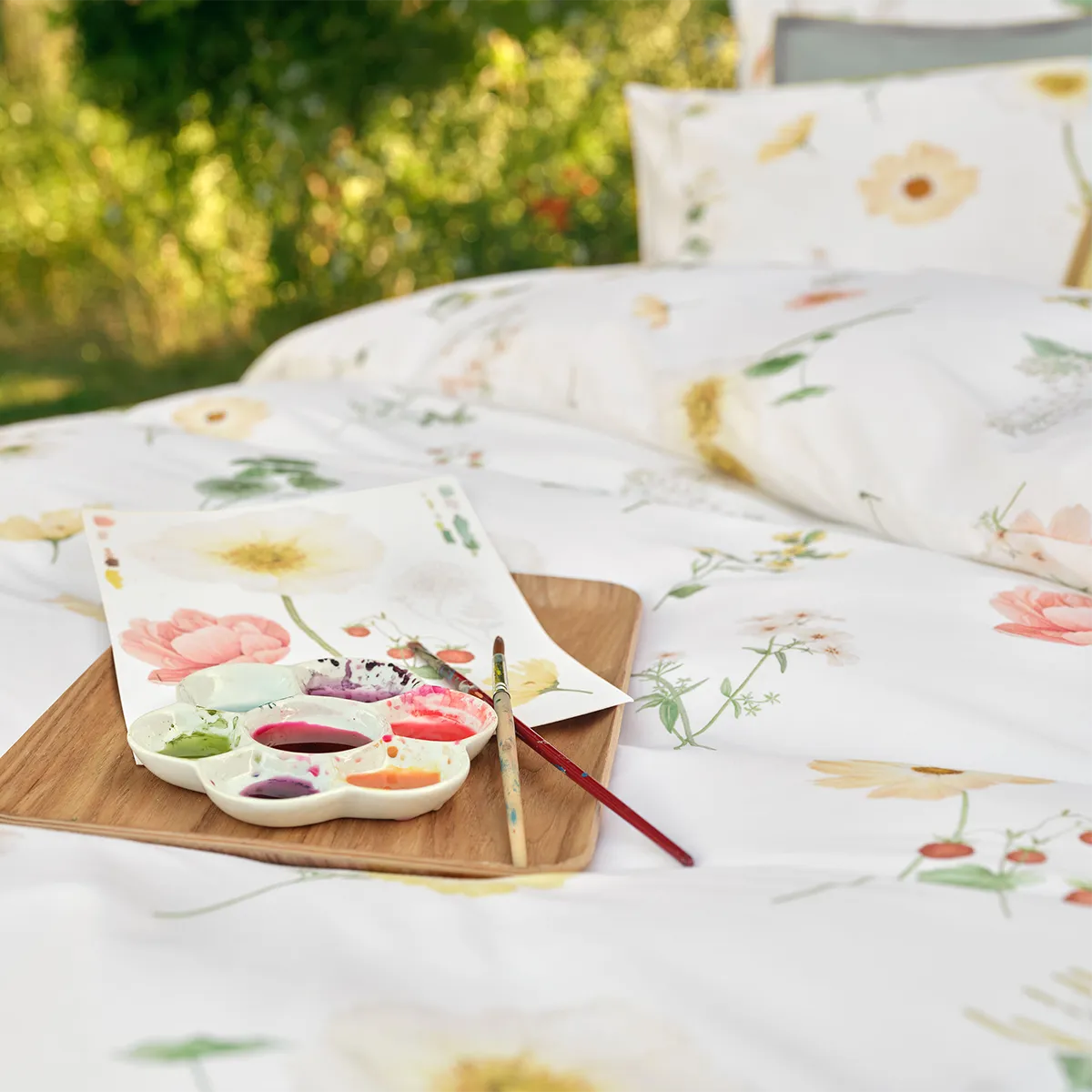 Schlossberg Bettwäsche Zinnia – Zeichnung und Kunstwerk Detailaufnahme der Schlossberg Bettwäsche Zinnia, auf der eine Wasserfarbenpalette und Pinsel auf einem Holzbrett abgebildet sind. Neben der Palette liegt ein gemaltes Blatt mit floralen Motiven, die den Blumen auf der Bettwäsche ähneln.