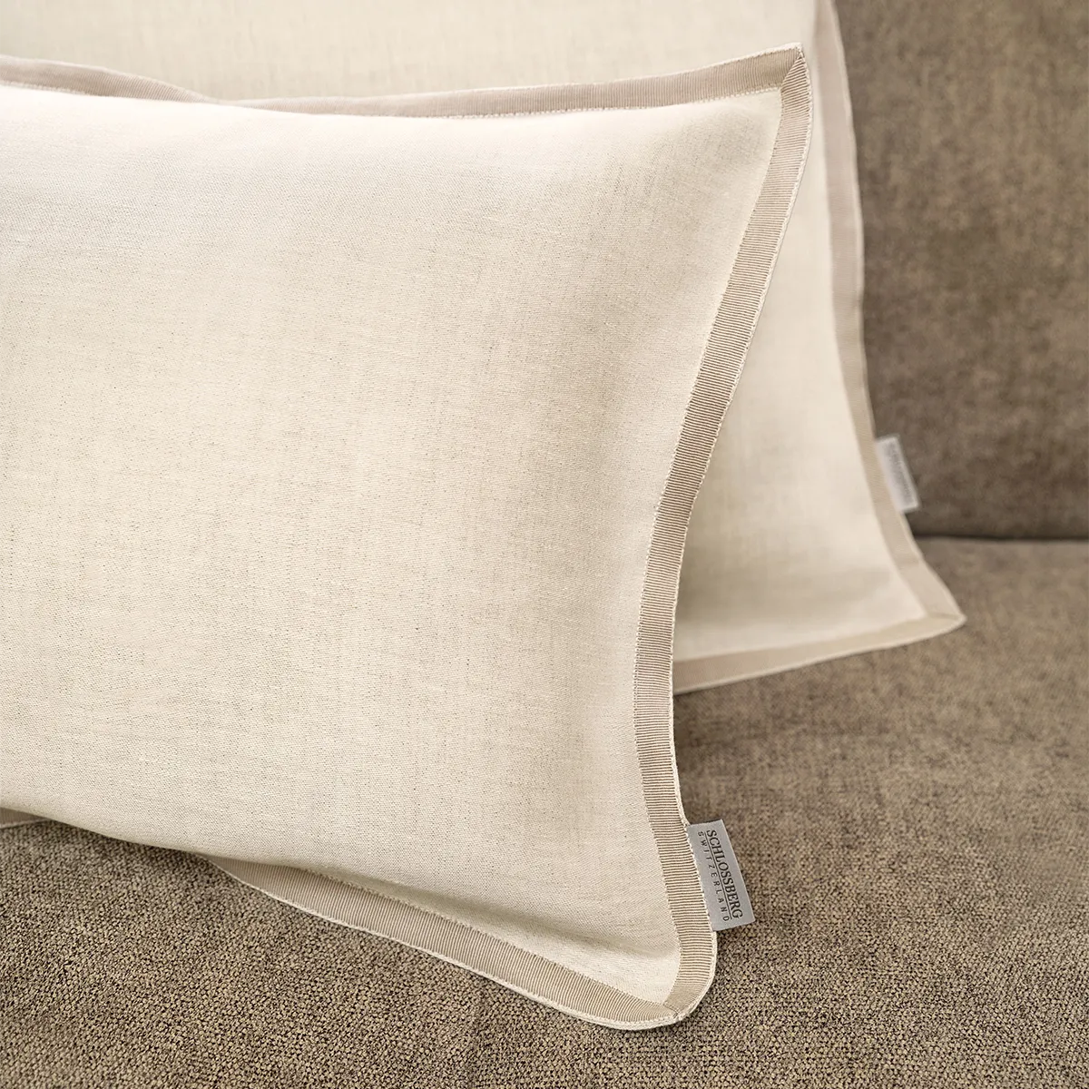 Detailaufnahme von zwei Schlossberg Zierkissen Solene Sand auf einem beigen Sofa. Die feine Struktur des Stoffes, kombiniert mit einer eleganten Paspelierung, verleiht den Kissen eine edle und natürliche Optik – perfekt für stilvolle Wohnräume.