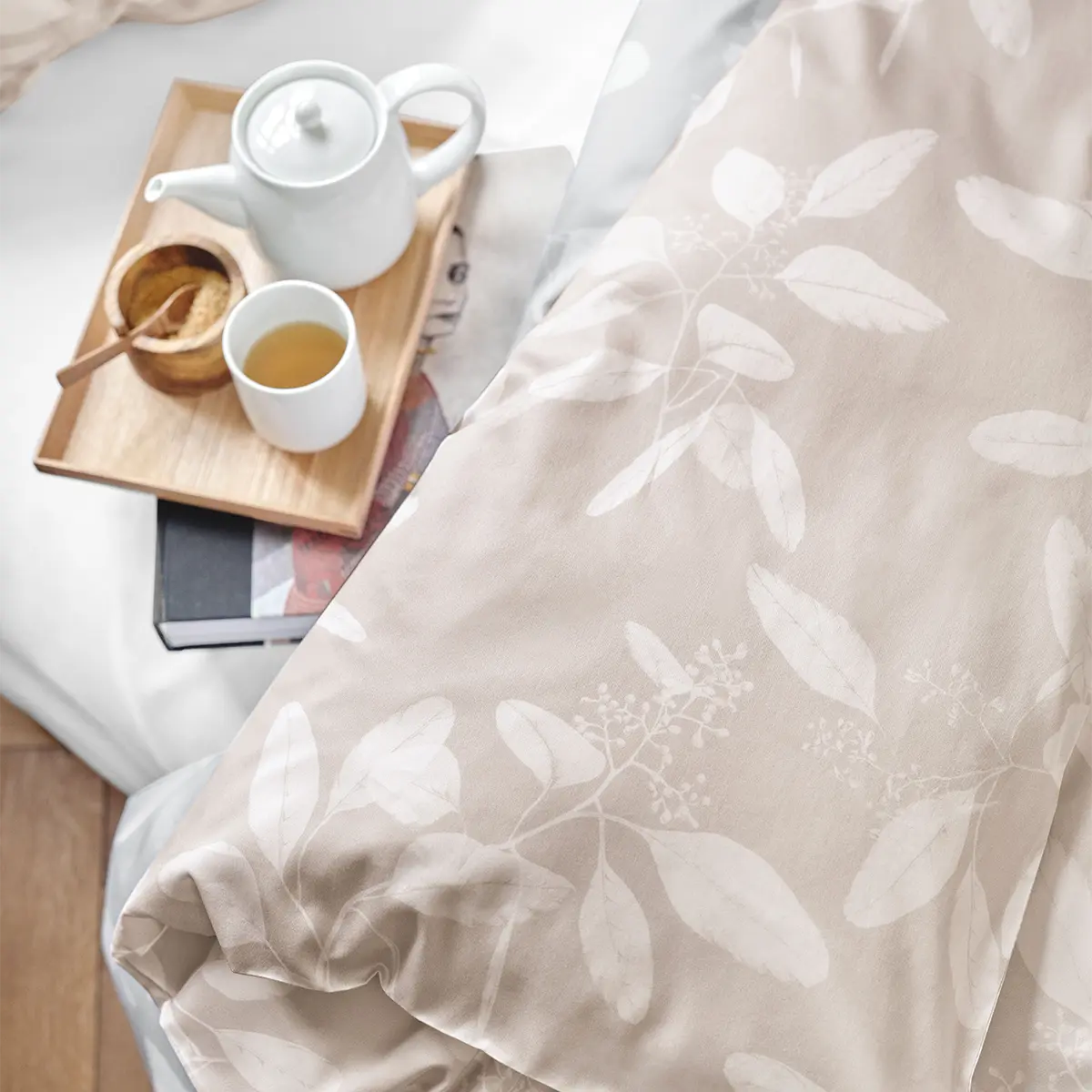 Eine Tasse Tee und eine Teekanne auf einem Tablett, arrangiert auf der beige-farbenen Omeo Bettwäsche mit Blättermuster.