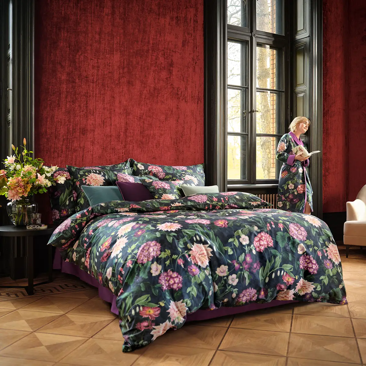 Ein grosszügiges Schlafzimmer mit hohen Fenstern und einer prächtigen, dunkelblauen Ophelia Bettwäsche, die ein florales Muster zeigt. Eine Frau steht am Fenster und liest ein Buch.