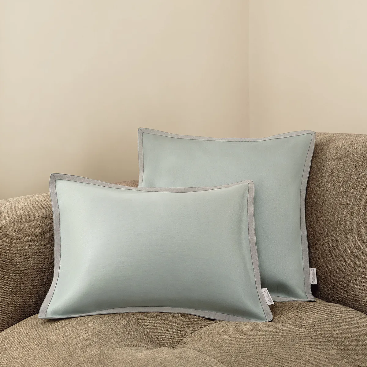 Zwei Schlossberg Zierkissen Solene in Sage auf einem beigen Sofa. Das edle Design in sanftem Salbeigrün mit kontrastierender Paspelierung sorgt für eine harmonische und stilvolle Wohnatmosphäre.