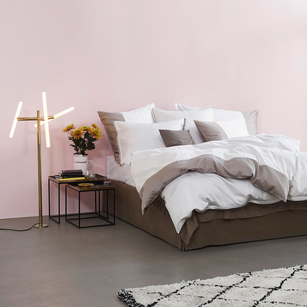 Modernes Schlafzimmer mit weisser und grauer Bettwäsche, Beistelltisch mit Blumen und Büchern, rosa Wand, grauem Boden und weissem Teppich.