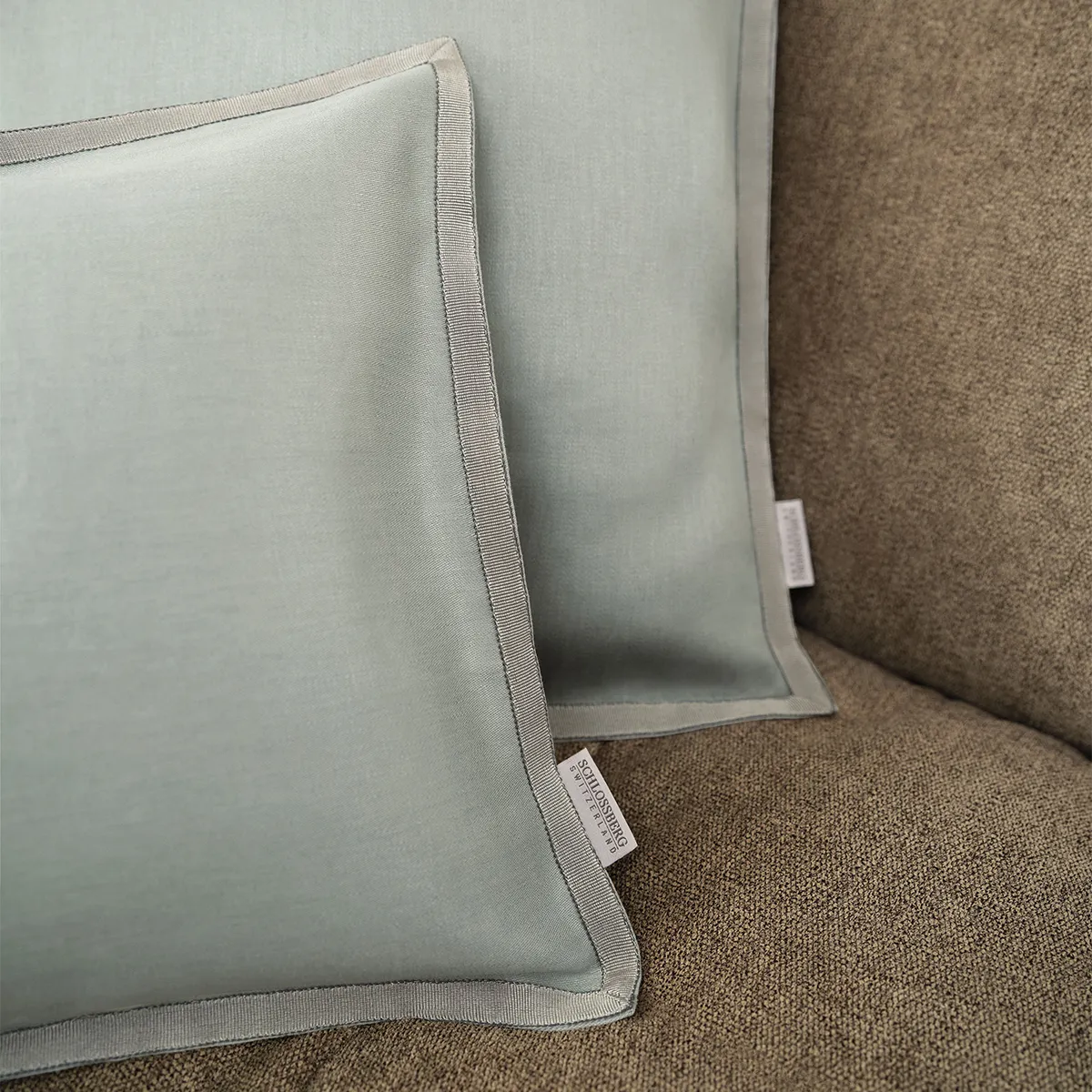Detailaufnahme von zwei Schlossberg Zierkissen Solene in Sage auf einem braunen Sofa. Die sanfte Salbeigrün-Färbung und die feine Paspelierung verleihen dem Kissen eine edle Note, ideal für eine stilvolle und gemütliche Einrichtung.