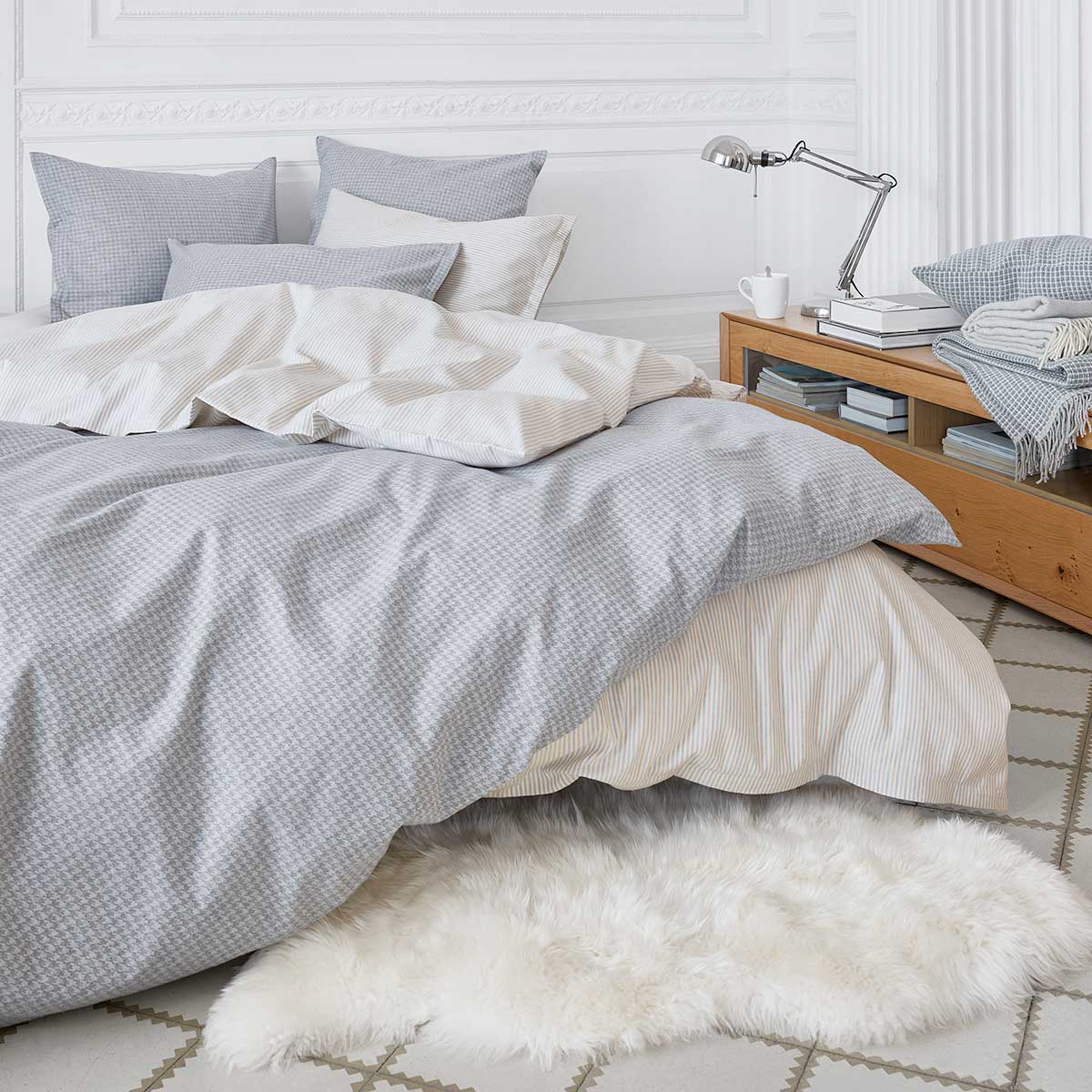Ein gemütliches Schlafzimmer dekoriert mit der Bettwäsche "Gustav" in einem feinen Streifenmuster, kombiniert mit grauen Akzenten, einem Holz-Nachttisch und einem Schaffellteppich.