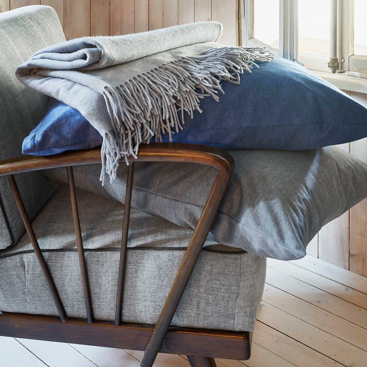 Caspar Kissen und Decke in dunkelgrau und blau, auf einem grauen Sessel mit grauer Decke.