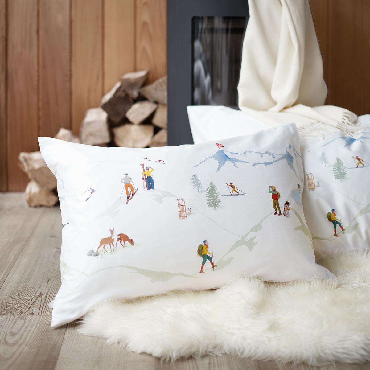 Kissen mit Alpina-Motiv Nahaufnahme eines Kissens mit alpinem Motiv vor einem Kaminofen, umgeben von Holzscheiten und einem weissen Fellteppich.