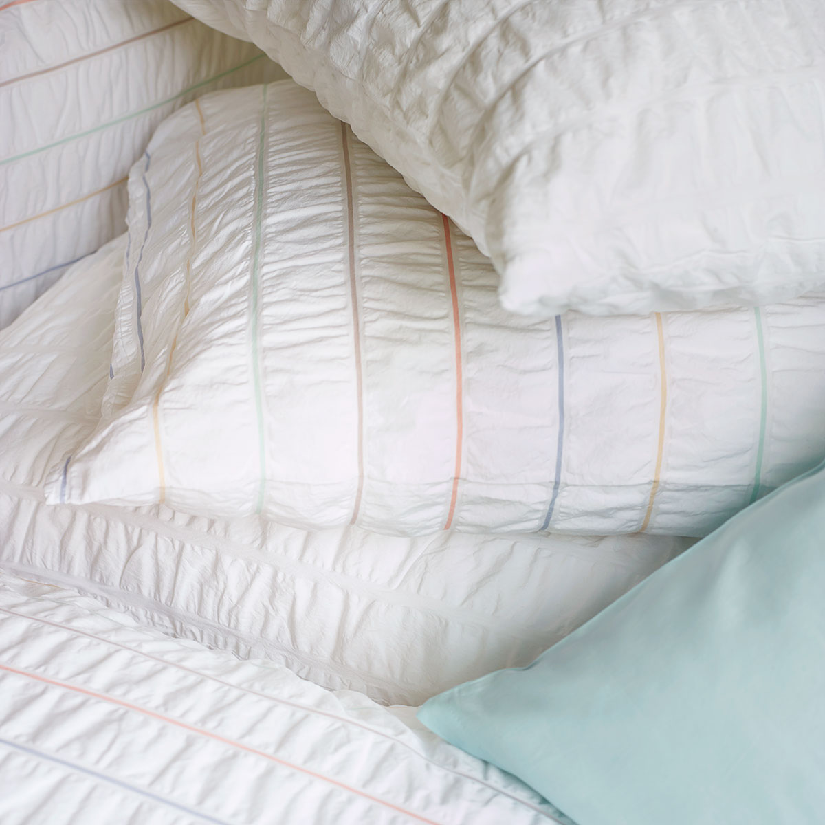 Detailansicht der Nelson Bettwäsche Nahaufnahme von Kissen und Duvet der Nelson Bettwäsche, die feine Streifen in Pastellfarben auf weissem Hintergrund zeigt.