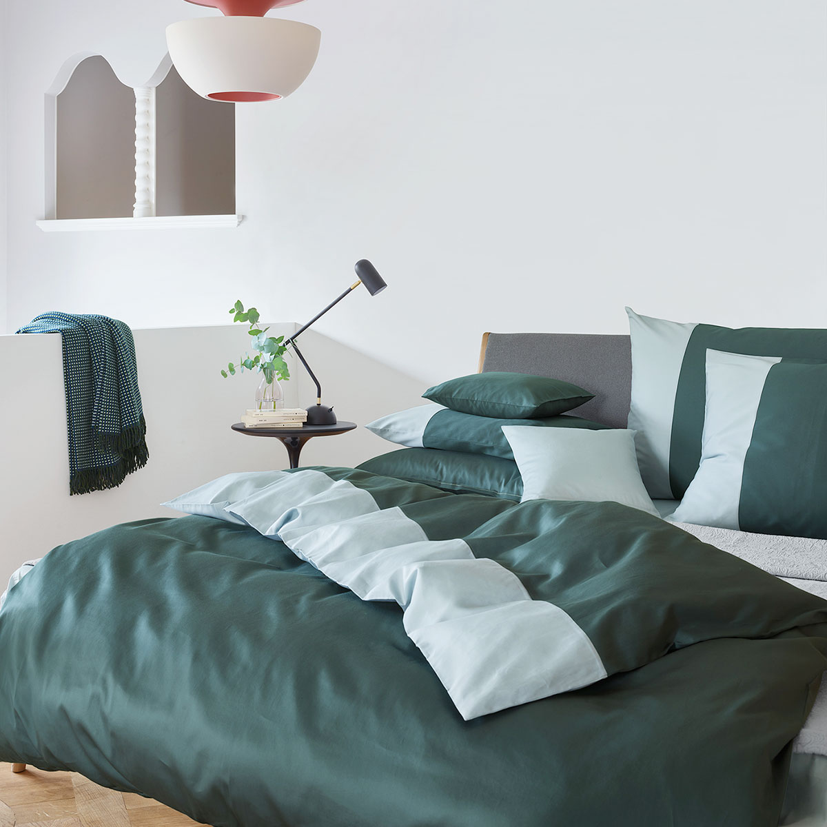 Schlafzimmer mit dunkelgrüner und hellblauer Bettwäsche, mehreren Kissen, Tisch mit Pflanze und Lampe, blau-weisser Decke im Hintergrund.