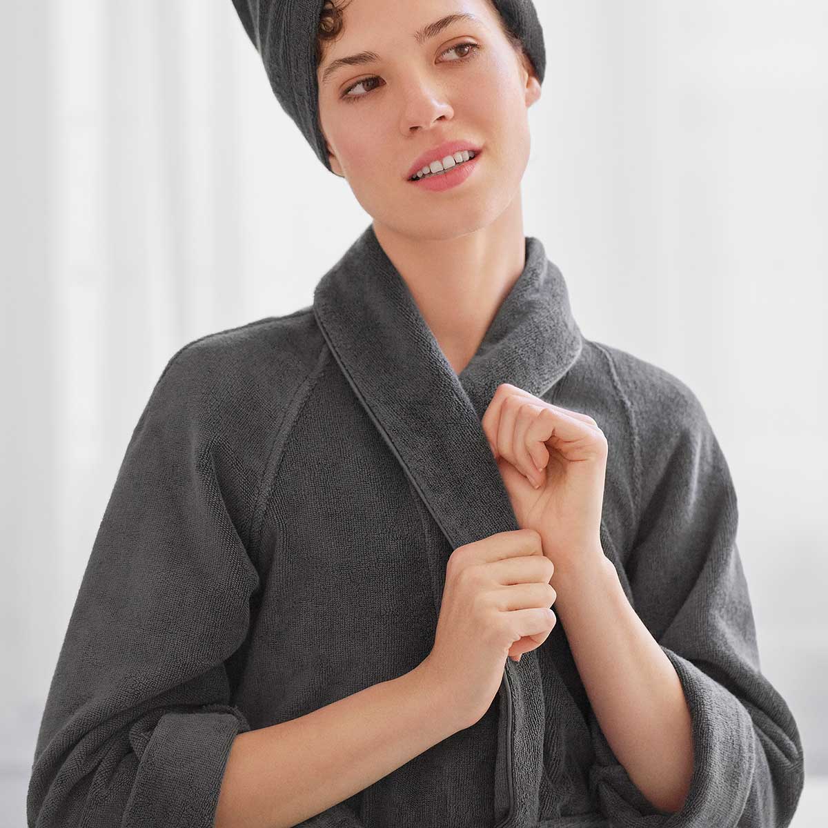Schlossberg Bademantel Coshmere Grau mit Haarturban Eine Frau trägt den grauen Schlossberg Bademantel aus der Coshmere-Kollektion und einen Haarturban, während sie den Bademantel schliesst.