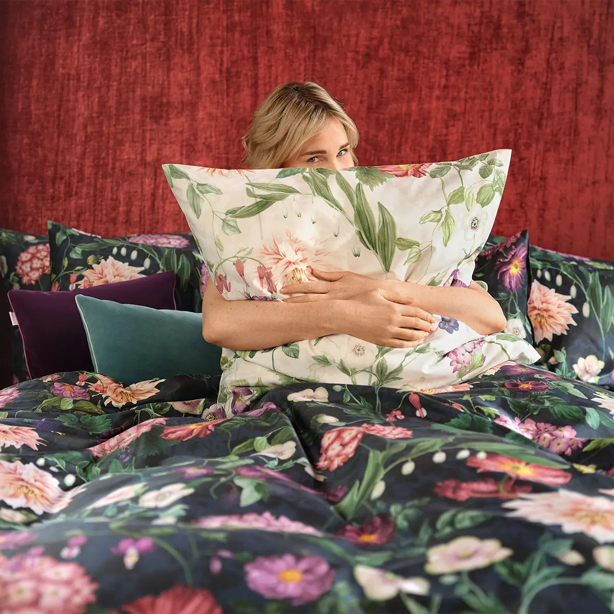 Eine Frau umarmt ein Kopfkissen mit dem floralen Design der Ophelia Bettwäsche. Der Hintergrund ist ein tiefroter Stoff, der eine gemütliche Atmosphäre schafft.