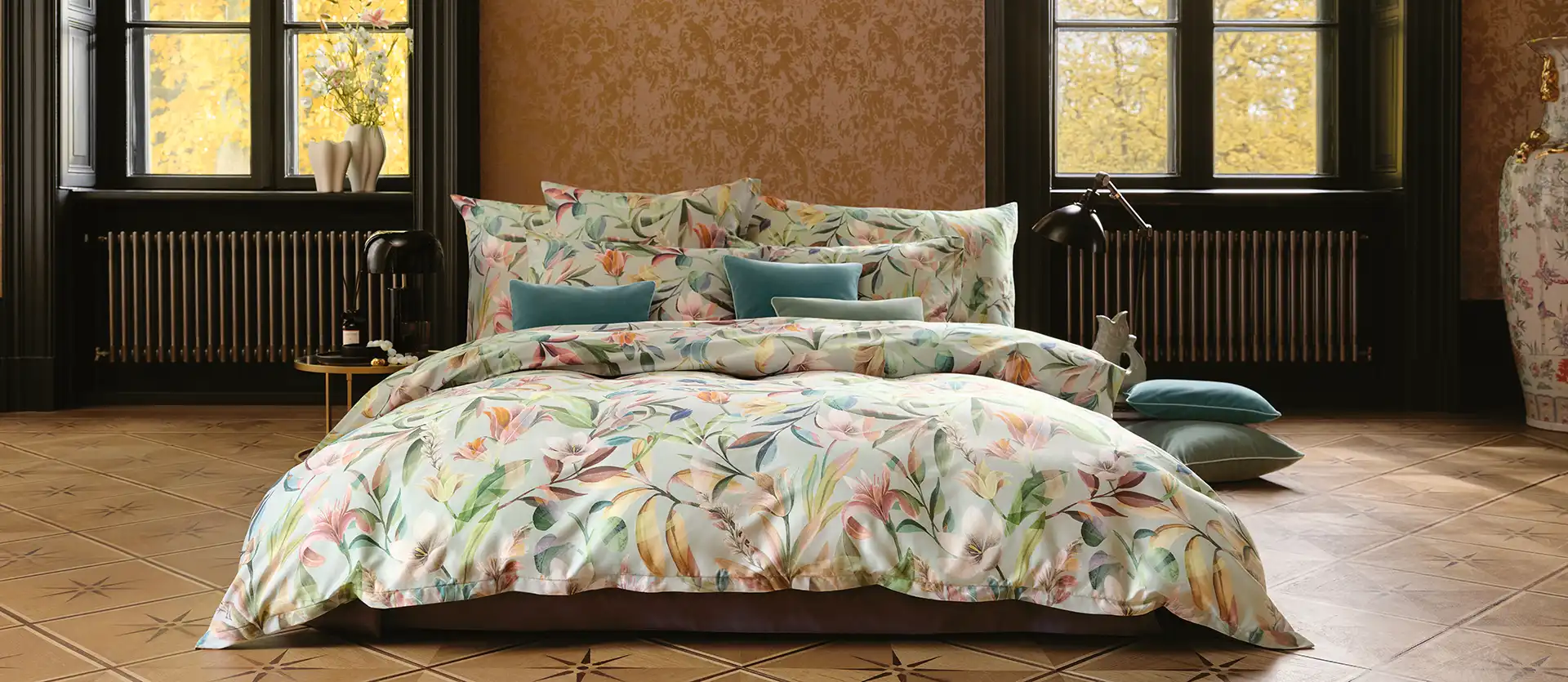 Ein Schlafzimmer mit Schlossberg Bettwäsche im Adele Design, das florale Muster in sanften Farben zeigt. Kissen in verschiedenen Größen und Farben sind auf dem Bett arrangiert, vor einem großen Fenster mit Herbstblick.