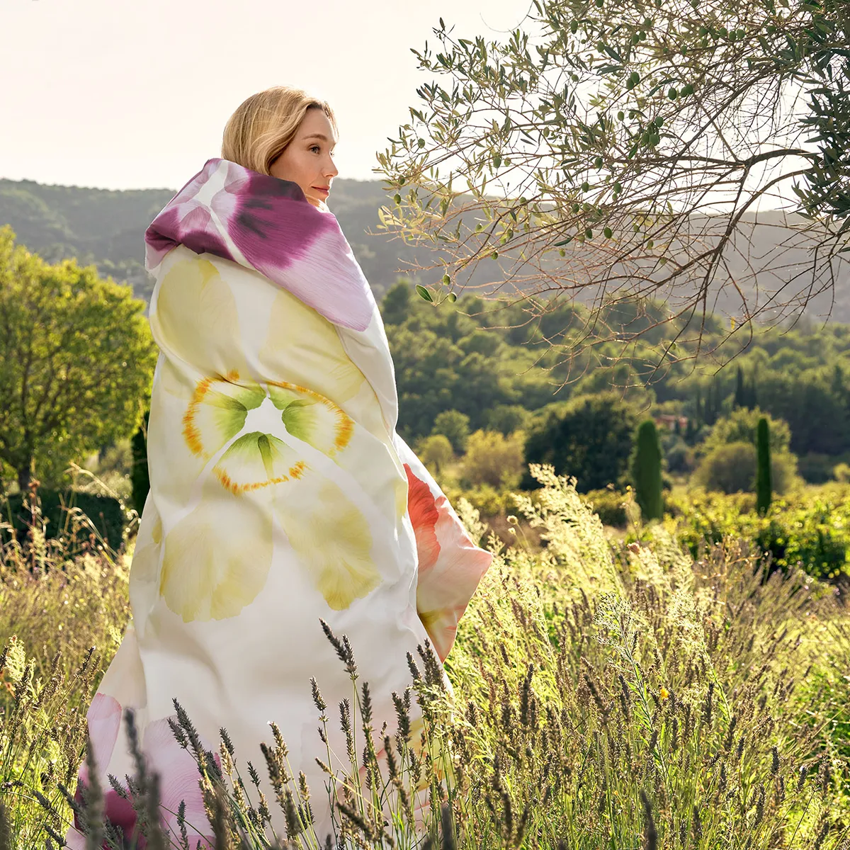 Eine blonde Frau steht inmitten einer sonnendurchfluteten Wiesenlandschaft, eingehüllt in die luxuriöse Schlossberg-Bettwäsche Aurora. Das weiche, mit floralen Blüten verzierte Design harmoniert perfekt mit der idyllischen Natur im Hintergrund.