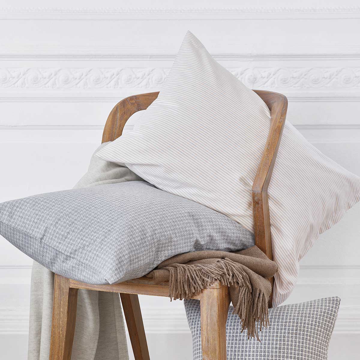 Kissen aus der Bettwäsche "Gustav", stilvoll auf einem Holzstuhl arrangiert, mit einer Mischung aus beige-grauen Streifen und einem dezenten Muster, dekoriert mit einer weichen Decke.