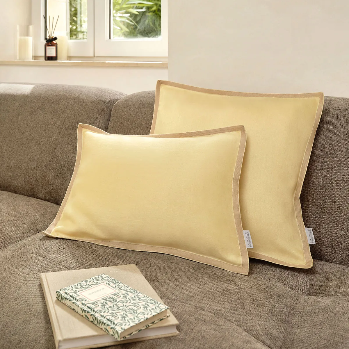 Schlossberg Zierkissen Solene Sun – Stilvolle Wärme für Ihr Zuhause Zwei Schlossberg Zierkissen Solene Sun in warmem Gelbton auf einem beigen Sofa, begleitet von zwei stilvollen Notizbüchern. Die elegante Paspelierung und die hochwertige Verarbeitung sorgen für eine harmonische und gemütliche Wohnatmosphäre.