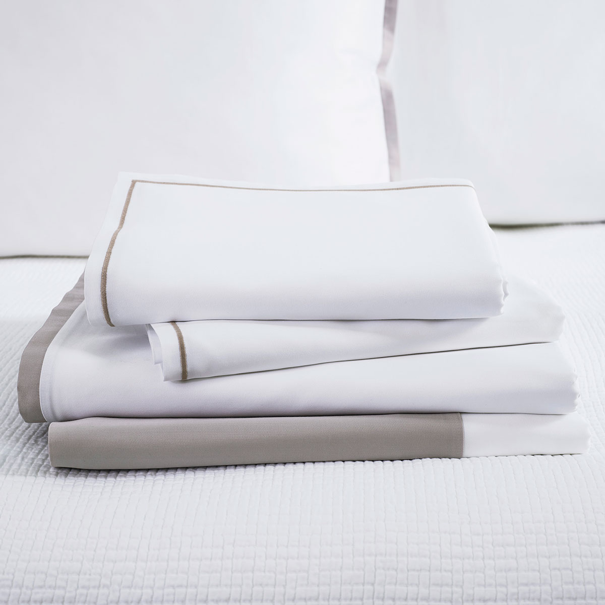 Gefaltete Bettwäsche Allen Ein Stapel weisser und grauer Bettwäsche liegt auf einem Bett mit weissem Bettbezug. Die Bettwäsche ist ordentlich gefaltet und zeigt dezente graue Akzente.