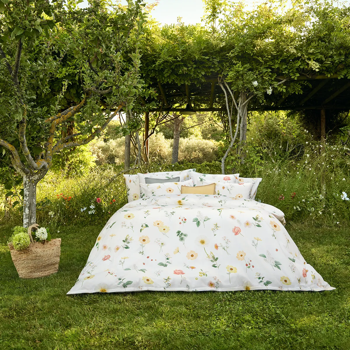 Schlossberg Bettwäsche Zinnia – Natürliches Design im Grünen Ein Bett mit Schlossberg Bettwäsche Zinnia steht mitten in einem idyllischen Garten unter einer grünen Pergola. Die Bettwäsche zeigt ein florales Design mit gelben, rosafarbenen und weißen Blüten sowie zarten grünen Blättern auf einem weißen Hintergrund.
