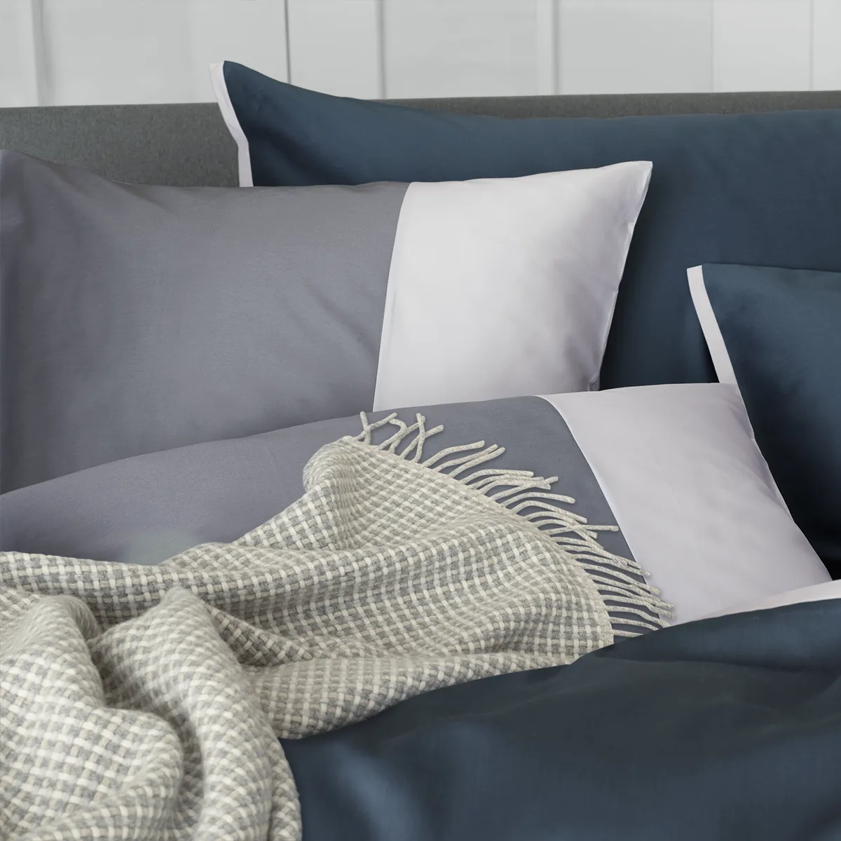 Ein modernes Bett mit Schlossberg Bettwäsche im Allen Design, das sich aus dunkelblauen und grauen Kissenbezügen zusammensetzt, begleitet von einem weichen, karierten Überwurf.