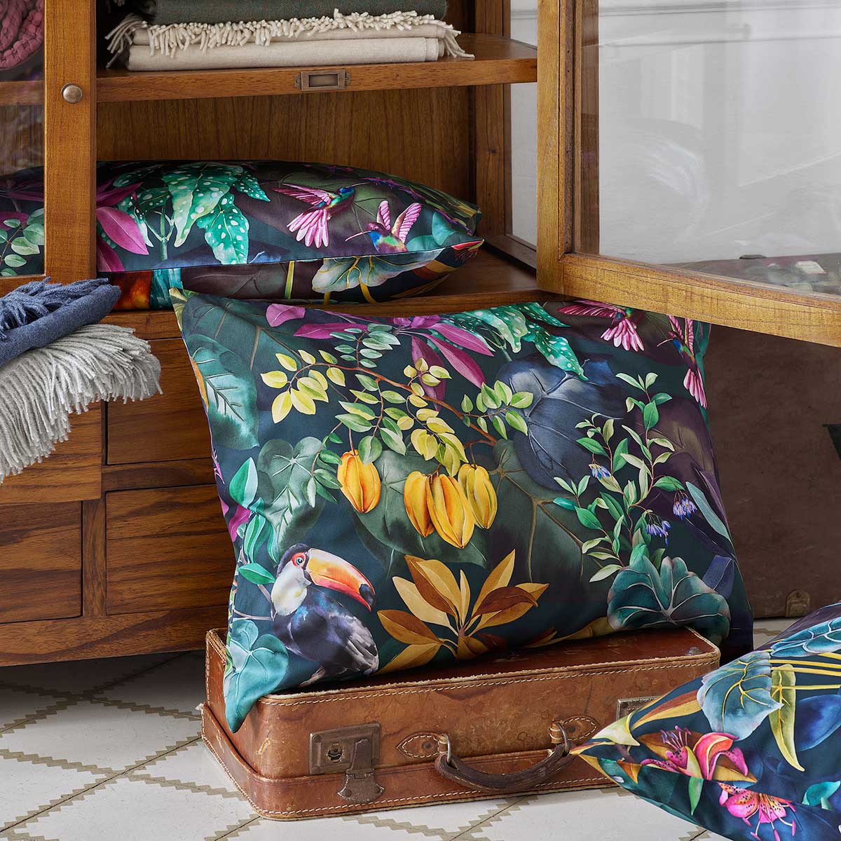 Exotische Kissen Jane mit Tropenmuster Kissen aus der Jane-Bettwäsche Kollektion mit exotischem Tropenmuster in kräftigen Farben, präsentiert auf einem Holzschrank.