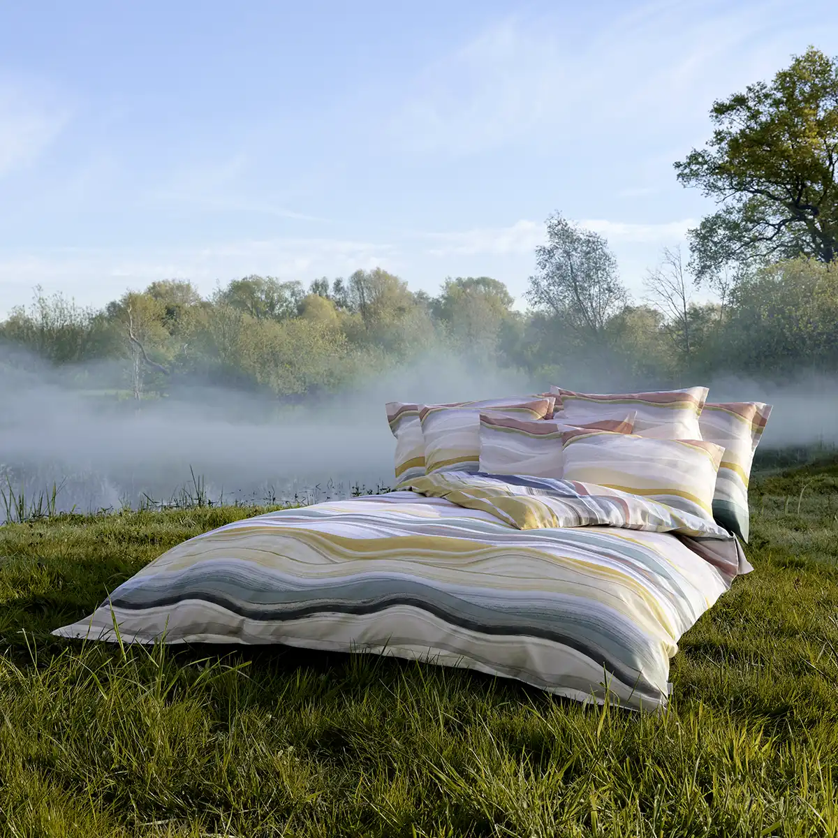 Linus Bettwäsche im Freien vor nebliger Landschaft Bett mit Linus Bettwäsche im Freien auf einer Wiese, umgeben von nebliger Landschaft bei Sonnenaufgang.