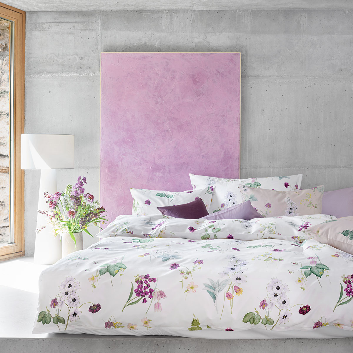 Schlafzimmer mit der Bettwäsche "Freya" und pastellfarbenen Akzenten Ein modernes Schlafzimmer mit weisser Bettwäsche "Freya", dekoriert mit floralen Mustern und pastellfarbenen Kissen, in einem Raum mit Betonwänden und einer rosa Wanddekoration.