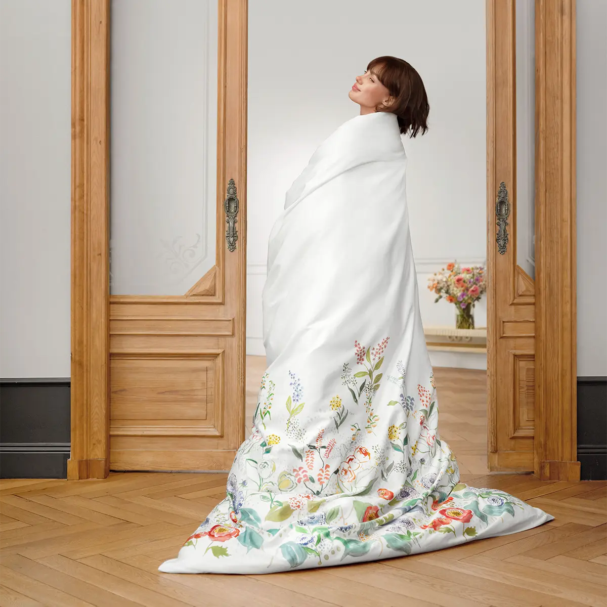 Bettwäsche Annika präsentiert Eine Frau in einem Raum, eingehüllt in die Bettwäsche Annika mit floralem Muster, vor einer hölzernen Tür.
